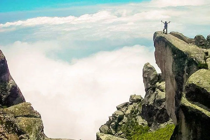 Ein Wanderer steht mit erhobenen Armen auf einem felsigen Gipfel über den Wolken.