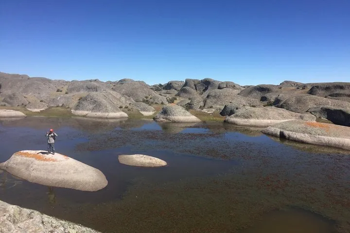 Un excursionista está de pie sobre una roca plana en un lago poco profundo en medio de un terreno rocoso.